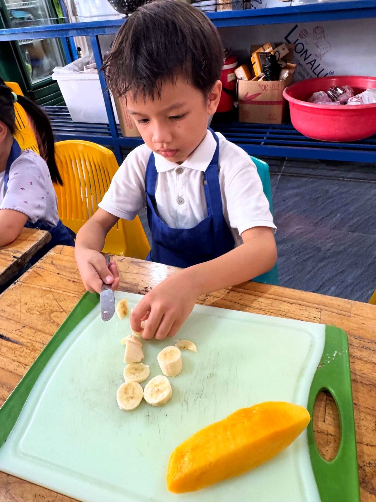 A Kindergarten child slicing banana beside half a mango in Cooking