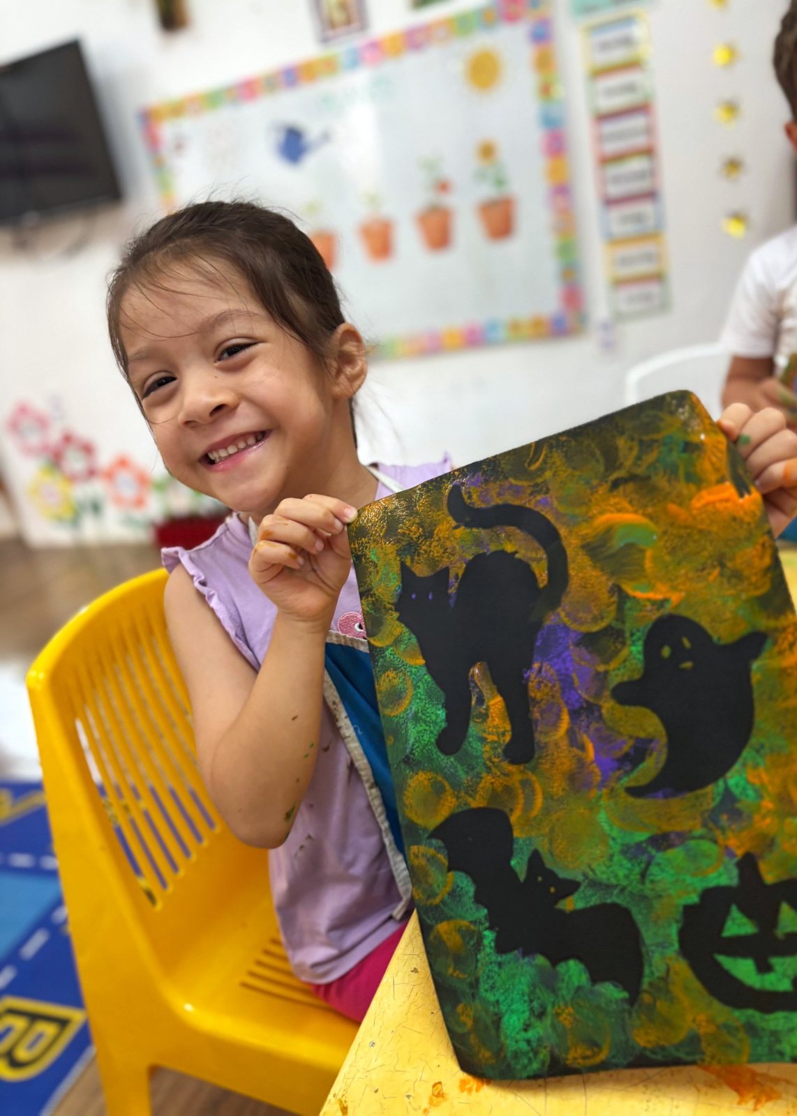 A Nursery child holding up her sponge-painted Halloween picture of cats and ghosts